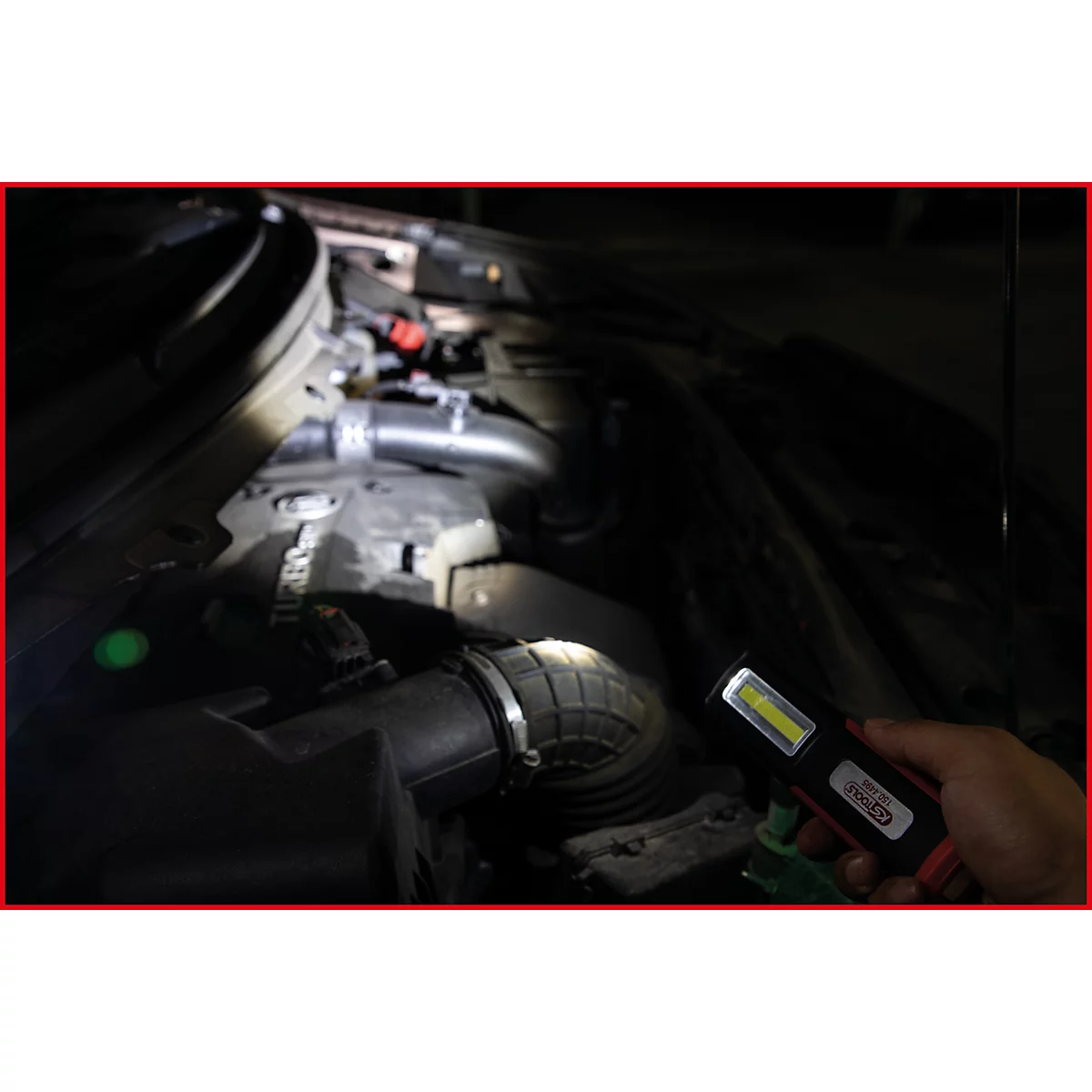 Een hand houdt een LED-werklamp in een donkere motorruimte. De lamp verlicht motoronderdelen.